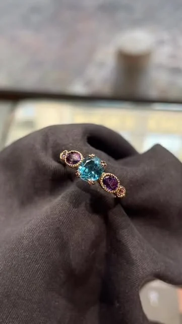 Bague à pierre centrale bleue et deux pierres violettes sur une étoffe sombre
